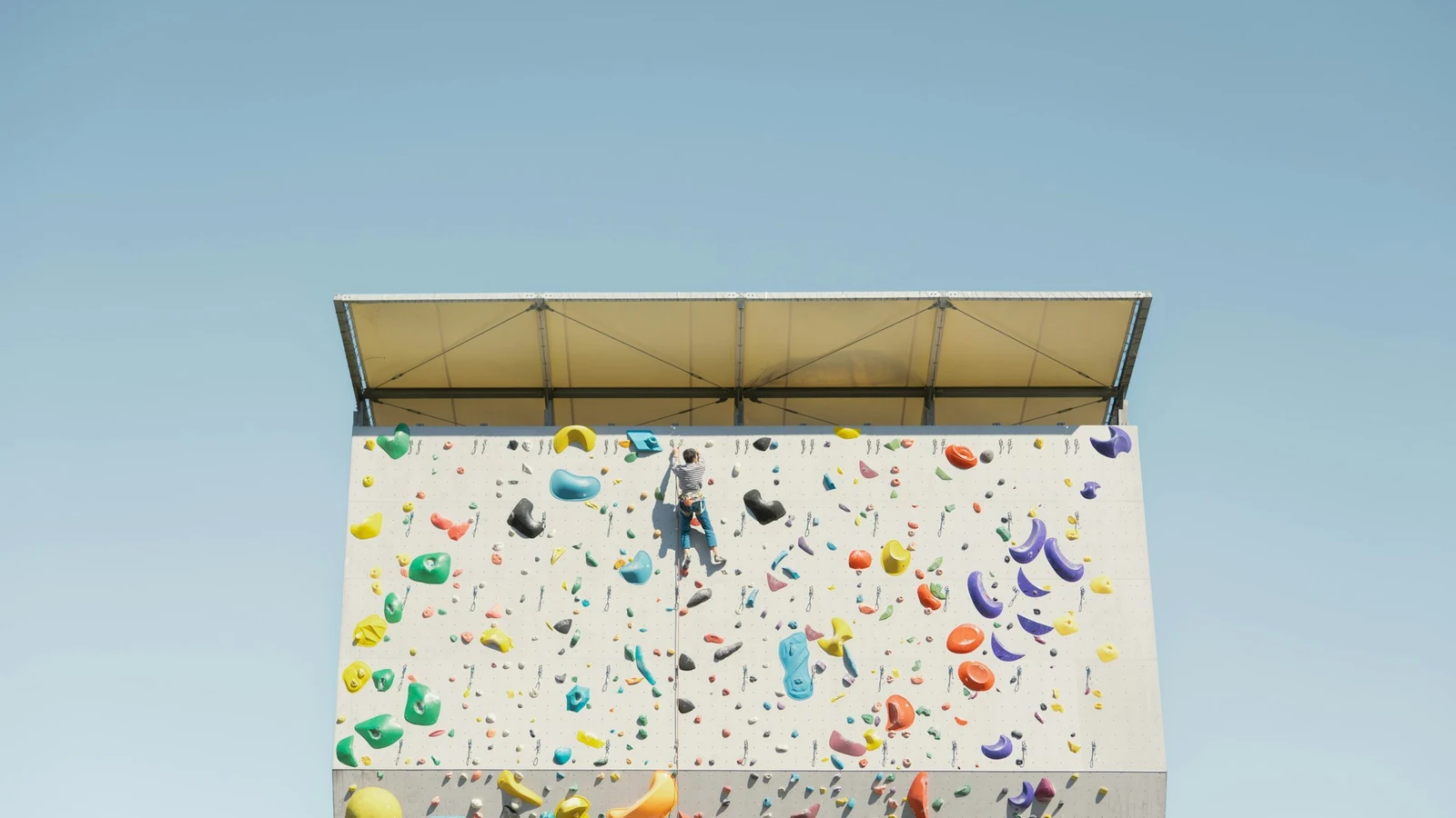 A lone climber partway up a tall outdoor climbing wall studded with brightly coloured holds, against a clear blue sky.